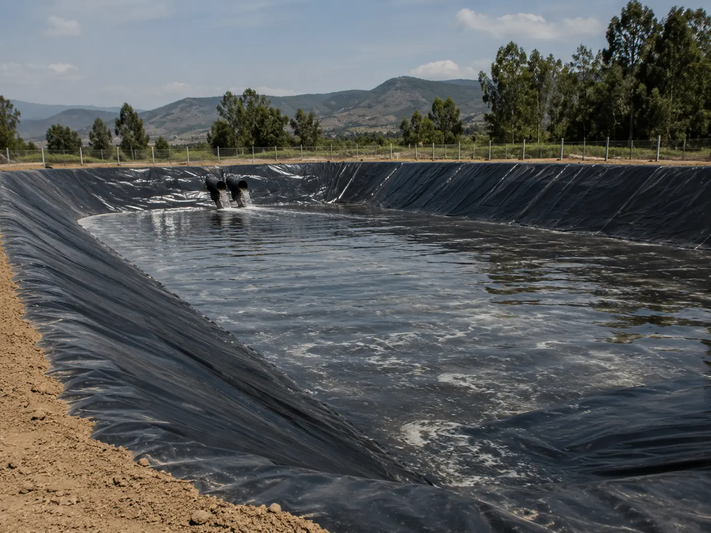 Laguna de tratamiento revestida con geomembrana, con dos tuberías de entrada descargando efluente y cerco perimetral